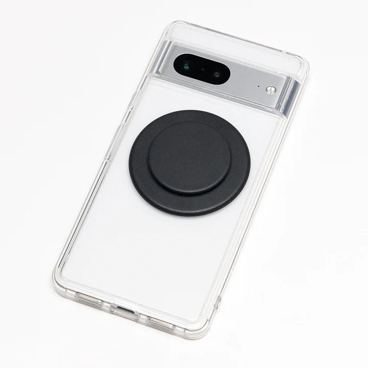 A smartphone with a clear case is shown lying flat with a PopGrip MagSafe in black attached to its back. The PopGrip features a round, flat surface with a matte black finish, positioned centrally on the phone's back. The phone's camera module is located near the top edge, and the clear case allows visibility of the phone's design, highlighting the PopGrip's sleek and modern appearance while providing a functional grip.