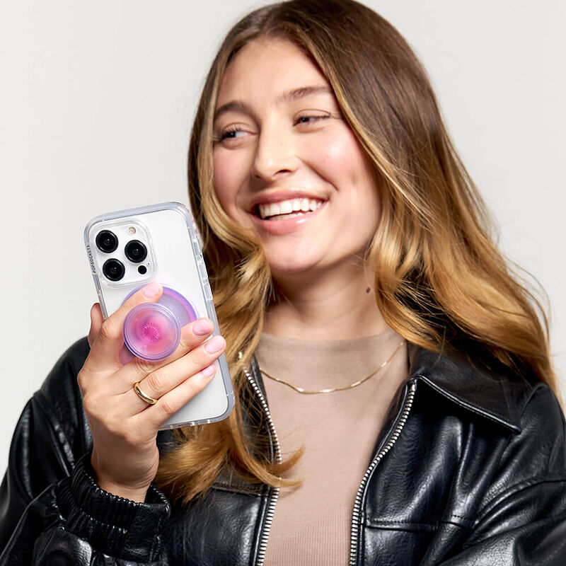 A woman with long, wavy hair and wearing a black leather jacket is smiling and holding a smartphone with a clear case. The phone has an Aura PopGrip for MagSafe attached to its back, featuring a gradient color pattern transitioning from pink in the center to purple on the edges. The woman is holding the phone with the PopGrip extended, showcasing its functionality and stylish design.