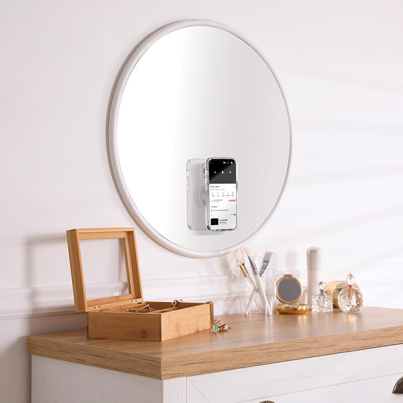 A round mirror is mounted on a wall above a wooden dresser. Attached to the mirror is a smartphone held by a PopSockets Suction Mount for MagSafe in the color horchata. The dresser has various items on it, including a wooden jewelry box, makeup brushes, a small tray with perfume bottles, and other decorative objects. The PopSockets mount securely holds the phone on the mirror's surface, showcasing its functionality and design.
