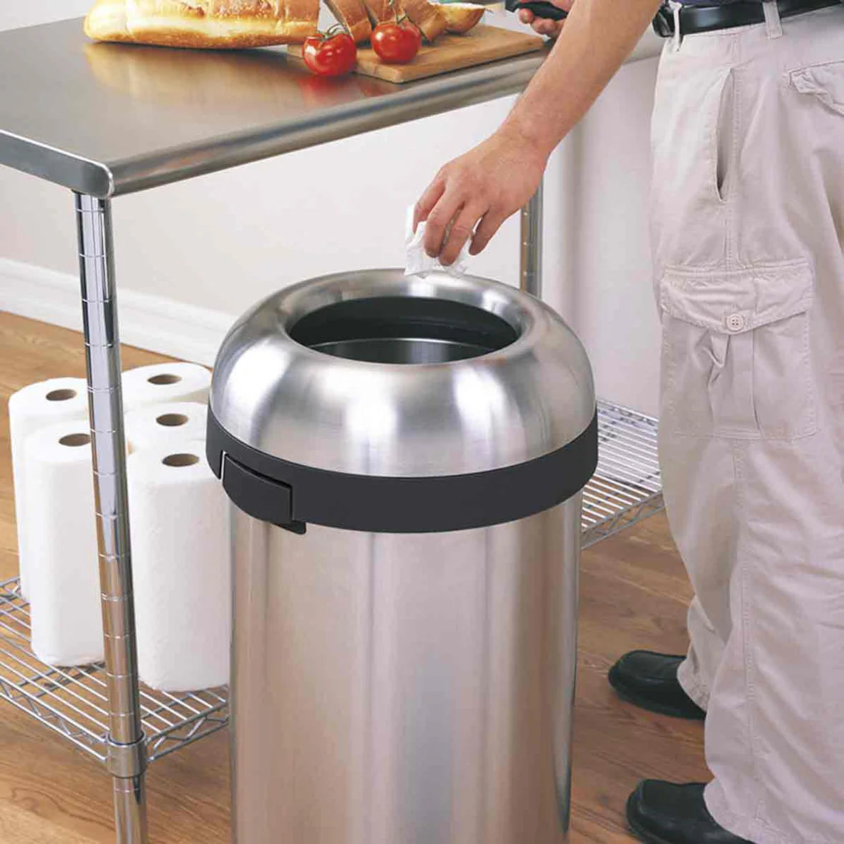 The image shows a person disposing of waste into a large, stainless steel trash can with a wide, circular opening at the top. The trash can has a sleek, modern design with a polished finish and a black band around the top edge. The setting appears to be a kitchen or food preparation area, as there is a metal countertop with tomatoes and paper towels on a nearby rack. The trash can's design is both functional and stylish, making it ideal for high-traffic areas like kitchens, cafeterias, or commercial spaces