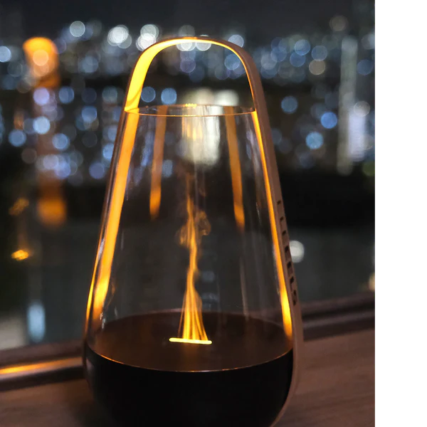 A modern flame-inspired ambient light with a sleek black base and a transparent glass enclosure, featuring a warm yellow-orange light resembling a flame. The light is placed on a wooden surface with a blurred cityscape of twinkling lights visible in the background, creating a cozy and elegant atmosphere. The integrated handle adds to its portability and aesthetic appeal.
