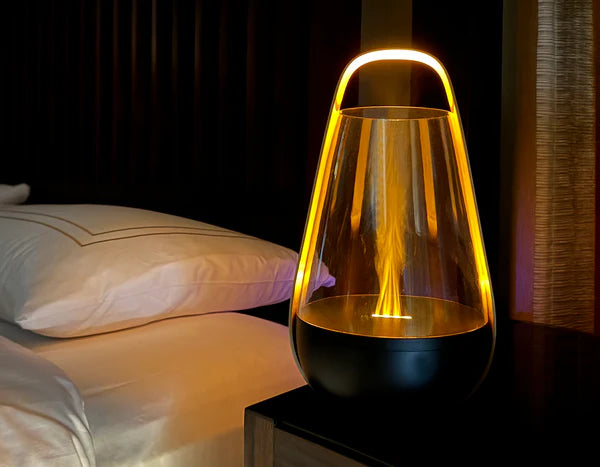 A sleek and modern flame-inspired ambient light placed on a bedside table. The device features a black base and a transparent glass enclosure with a warm yellow-orange light resembling a flame. Its minimalistic design and soft glow create a cozy and relaxing atmosphere, complementing the bedroom setting with white pillows and dark wooden furniture.