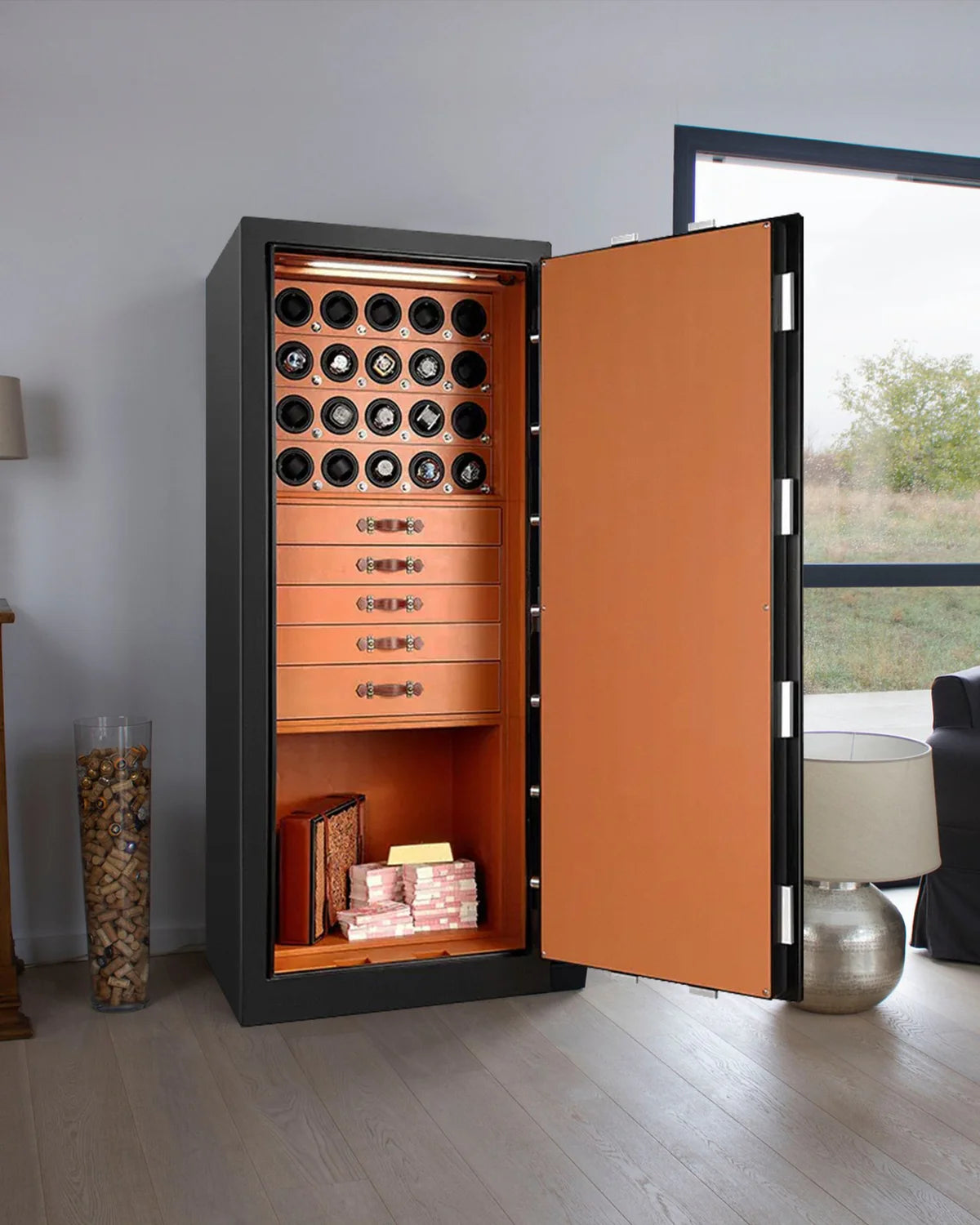 A large, open safe with a black exterior and a vibrant orange interior situated in a modern, well-lit room with a large window. The top section features sixteen watch winders. Below the watch winders are four drawers with decorative handles for organized storage.
