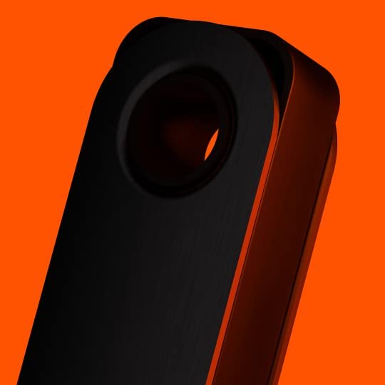 The image is a close-up shot of the Ledger Nano S Plus hardware wallet, focusing on the upper part of the device. The device is shown against a bright orange background, which contrasts sharply with the black, metallic surface of the Ledger Nano S Plus. The image highlights the circular hole at the top of the device, typically used for attaching a keychain or lanyard. The close-up view emphasizes the sleek and refined design, with the black color giving it a modern, high-tech appearance.