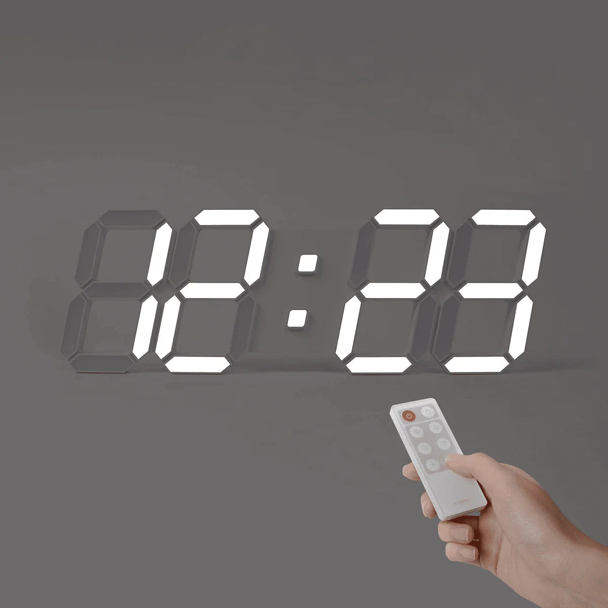 A digital wall clock with large, bright illuminated numbers displaying "12:88" is mounted on a gray wall. A hand holding a remote control is aimed at the clock, indicating the use of the remote to set or adjust the clock. The clock has a modern, minimalist design with easily readable numbers.