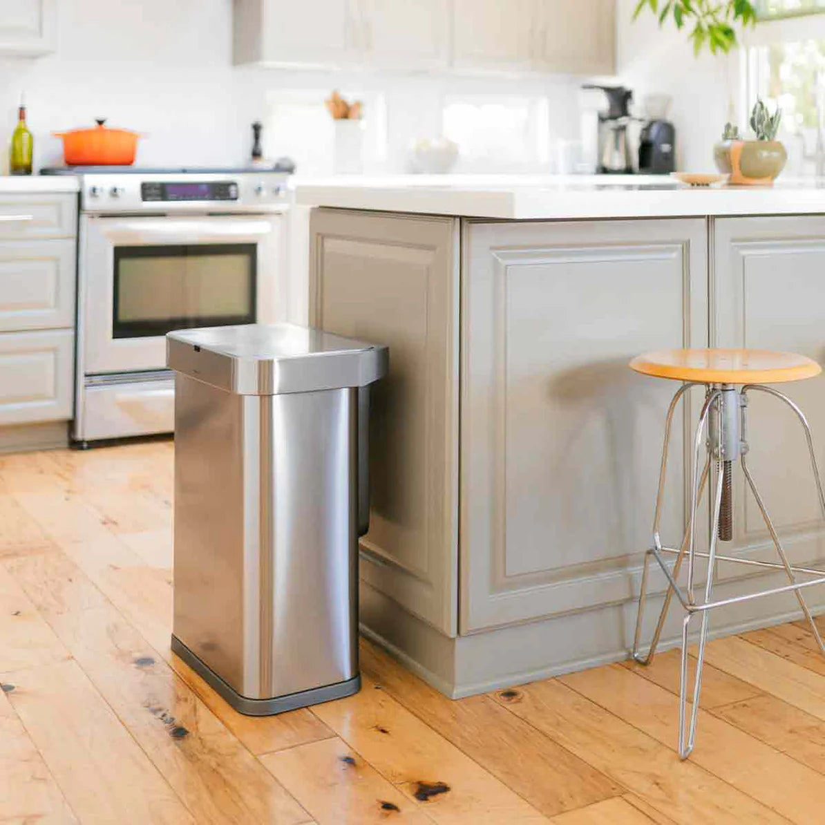 The image shows a bright and inviting kitchen with a Simplehuman stainless steel trash can positioned next to a kitchen island. The kitchen features light-colored cabinetry and a matching island with decorative molding. The stainless steel trash can blends seamlessly into the modern design, adding both functionality and a touch of elegance to the space.