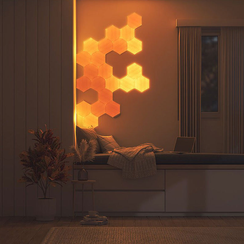 This image showcases a cozy bedroom setting with modern decor. A beige upholstered bed with plush pillows and a textured throw blanket occupies the right side of the room. On the wall above the bed, there's a stunning geometric light installation with hexagonal wooden panels that glow warmly, adding an artistic and ambient touch. The room features natural tones and minimalistic furniture, with a small wooden nightstand holding a plant and a clock, and a potted green plant in the corner, contributing to the