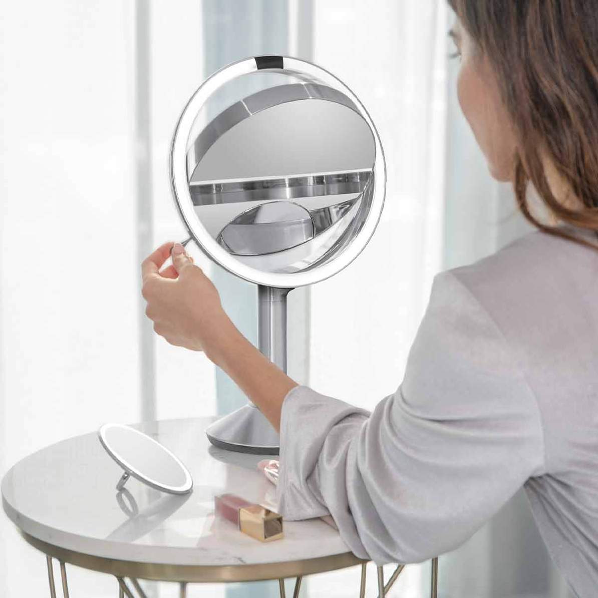 This image features the **Simplehuman sensor mirror** in use. The mirror's **bright LED ring light** provides excellent lighting for detailed tasks such as makeup application or grooming. The user is adjusting the mirror to their preferred angle, showcasing its **flexible tilt and rotation capabilities**. The mirror on the side table adds convenience, indicating the portability and sleek design of Simplehuman's product line. The **minimalist aesthetic** of both the mirror and the surrounding setup reinforce