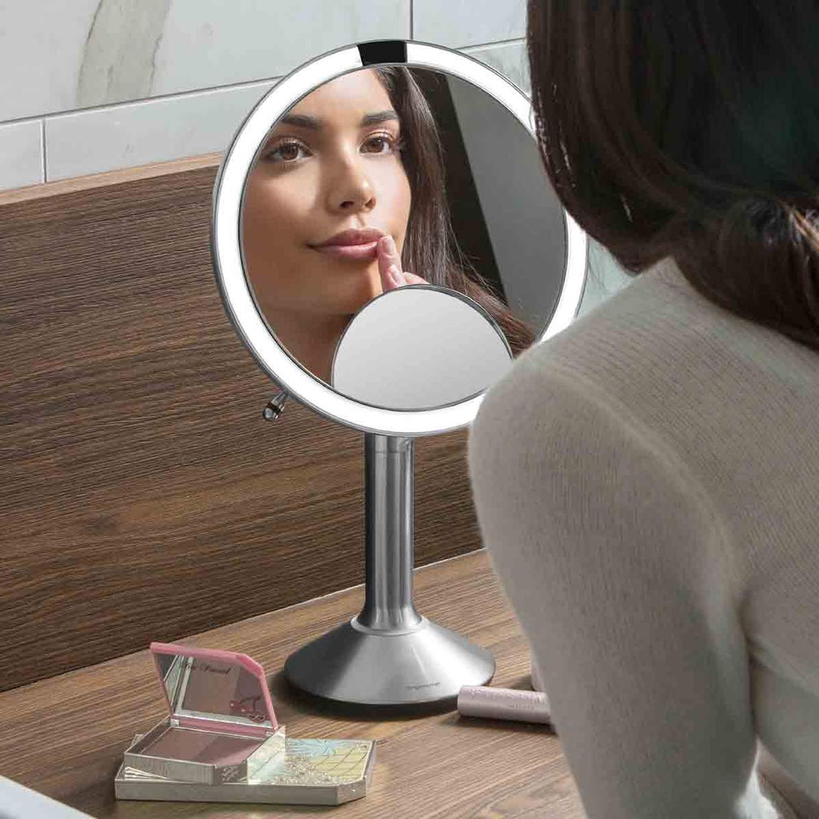 This image shows a person using the **Simplehuman sensor mirror** for makeup application. The **bright LED ring light** surrounding the mirror provides clear and bright illumination, ensuring precise visibility for tasks like applying lipstick. The design of the mirror allows for close-up detail work, supported by the built-in **magnifying mirror** seen at the bottom of the larger mirror. The mirror's **sleek, stainless steel** finish adds a modern touch to the surrounding environment, reinforcing its premi