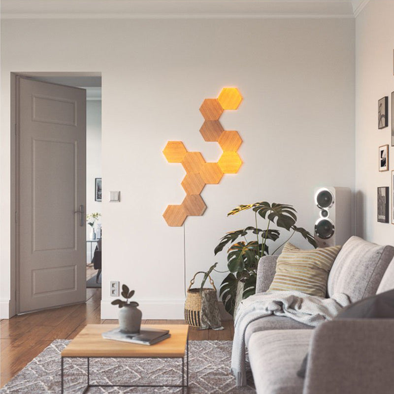This image depicts a modern, minimalist living room with a cozy, neutral-toned aesthetic. The focus is on a hexagonal wooden light installation on the wall, creating a warm, inviting ambiance with its soft glow. The room features a light gray sofa adorned with striped pillows and a throw blanket, and there's a small coffee table in the center with a simple plant and some reading material. In the background, a stylish plant adds a touch of greenery, while a tall speaker system sits by the wall, blending with