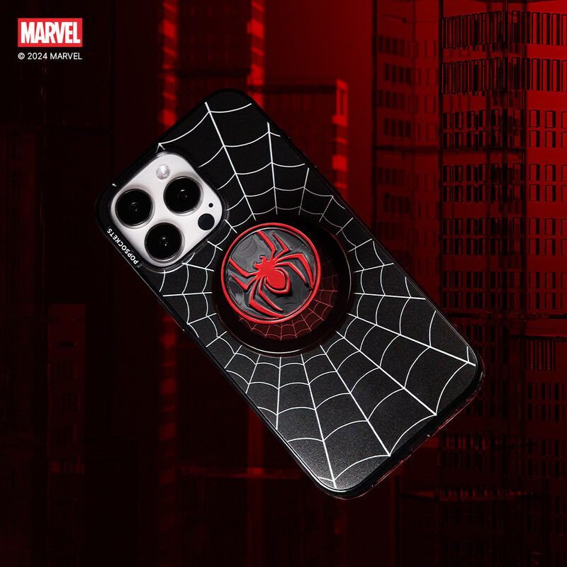 A smartphone with a black case featuring a white web pattern and a PopSockets Enamel Miles Morales Suit PopGrip for MagSafe. The PopGrip has a red and black design with the Miles Morales Spider-Man emblem in the center. The phone is angled against a dark red background with a cityscape pattern, highlighting the Spider-Man theme.