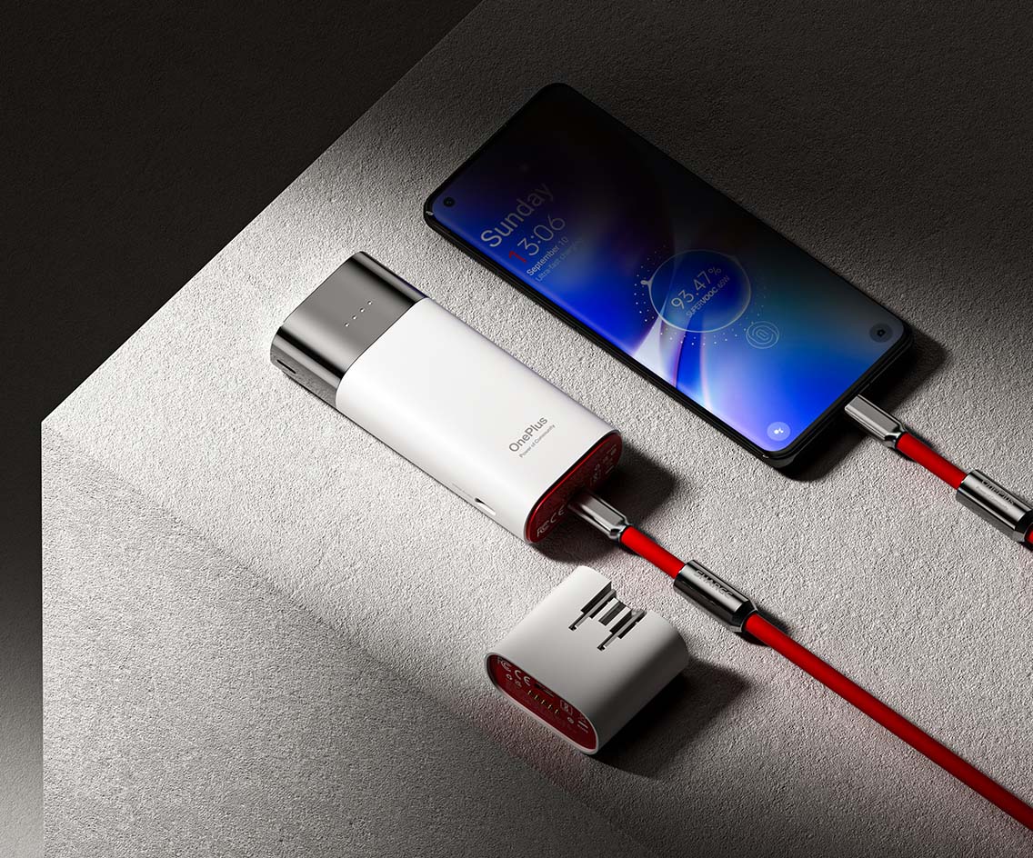 The image shows a sleek OnePlus charging device connected to a smartphone via a vibrant red cable. The device, branded with "OnePlus," is actively charging the phone, as indicated by the phone's screen displaying a 93% charge level. The charging setup is modern and stylish, with the charger featuring a metallic finish and a detachable component lying next to it, possibly an additional adapter or connector. The overall composition emphasizes the high-tech, premium quality of the charging device.