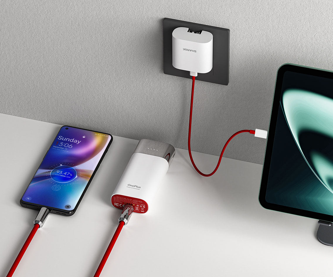 The image shows a charging setup featuring a OnePlus-branded charger connected to both a smartphone and a tablet. The smartphone is placed on a flat surface, showing a charging status of 93% with a time of 3:06 displayed on the screen. The charger is connected to a power outlet via a red cable, which also links to a tablet placed nearby. The power adapter in the wall outlet is branded with "Sharge," indicating a collaborative product between OnePlus and Sharge. The setup emphasizes a clean and efficient cha