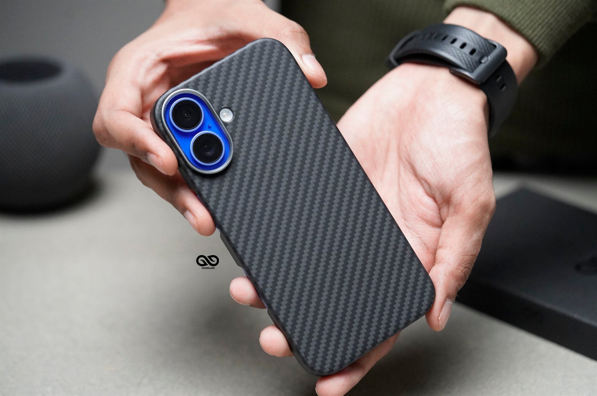 This image shows a person holding a sleek carbon fiber-textured phone case designed for a smartphone with a dual-lens camera setup, possibly an iPhone model. The case has a subtle, high-quality carbon fiber finish that provides a premium look and likely enhances grip and protection for the device. The black case contrasts elegantly with the blue ring around the camera lenses, adding a touch of style to the overall design. This case is ideal for users seeking both durability and a modern aesthetic.