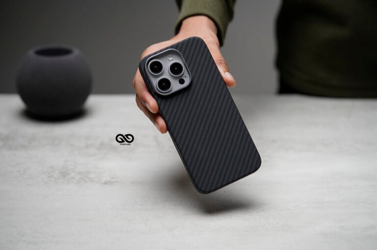 This image displays a person holding a phone case with a sleek, carbon fiber finish, designed for a smartphone featuring a triple-lens camera setup, likely a high-end model such as an iPhone Pro series. The carbon fiber texture on the case provides a sophisticated look and likely enhances grip and durability. The minimalist and modern design makes it ideal for users seeking both style and robust protection for their device.