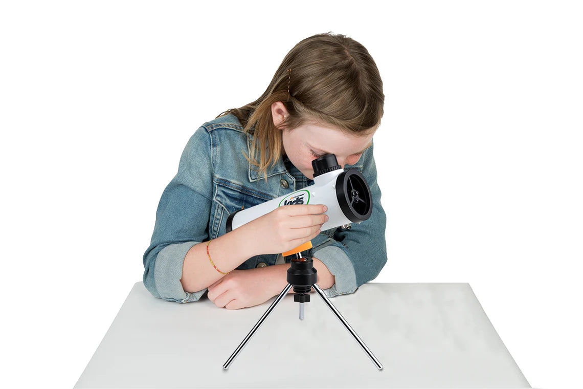 A young girl with light brown hair wearing a denim jacket is using a Celestron Kids STEM 50mm Newtonian Telescope. She is leaning forward and peering through the eyepiece of the white telescope, which is mounted on a small tripod and placed on a white table.