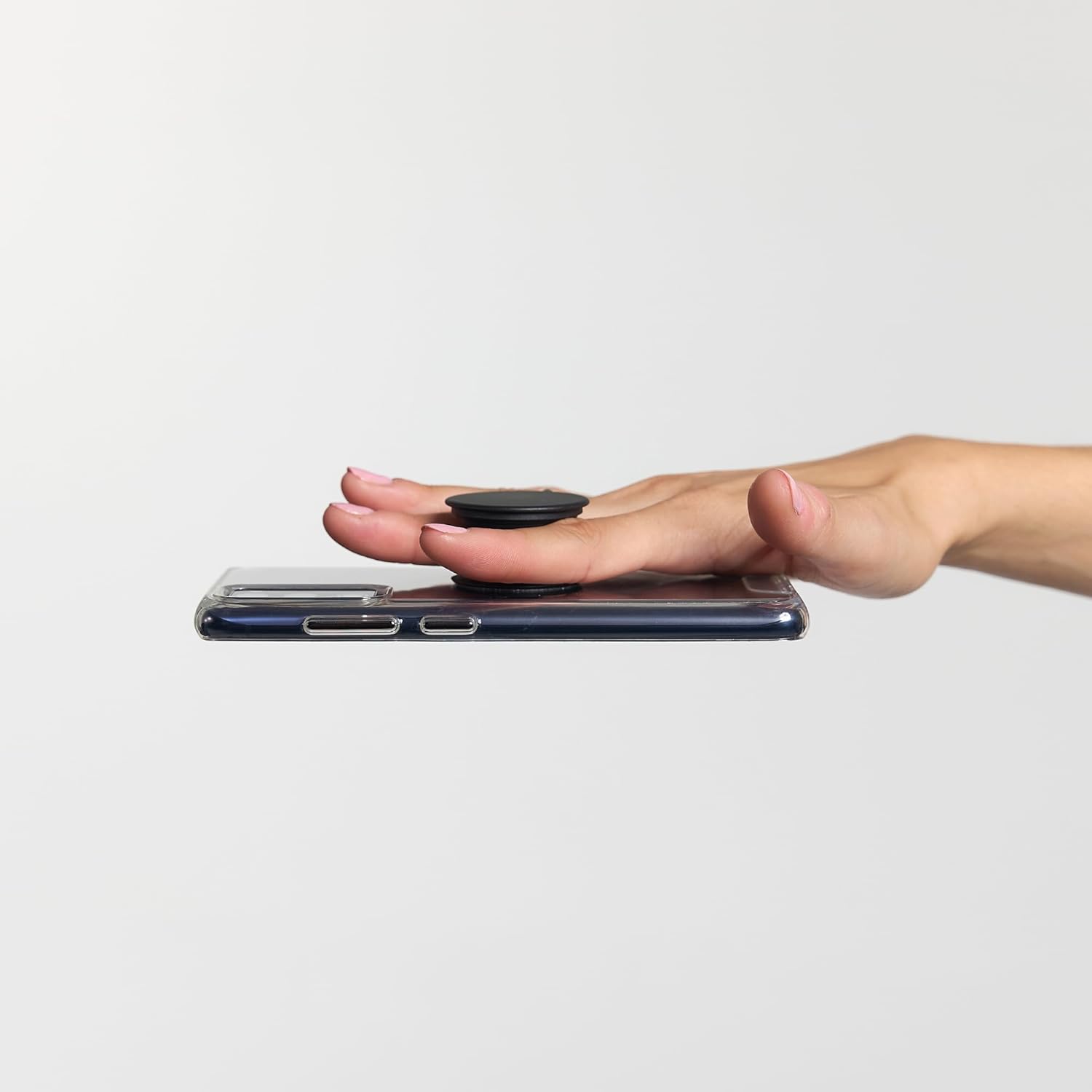  A person's hand holding a smartphone with a black PopSockets Phone Grip attached to the back. The phone is balanced on the person's fingers, demonstrating the secure and convenient hold provided by the PopSocket. The grip is in its collapsed state, providing a low-profile and ergonomic way to hold the phone. The background is plain, focusing on the hand and phone.