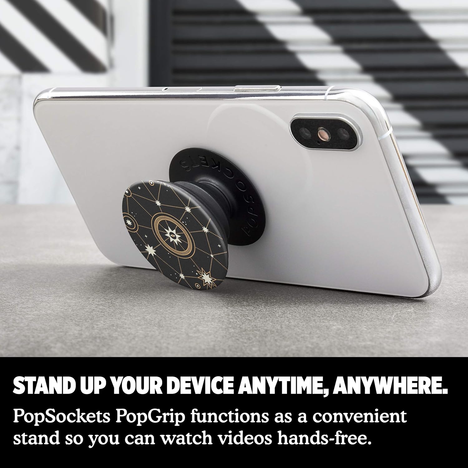 A white smartphone is propped up using a PopSockets grip with a star chart design, demonstrating its use as a stand. The text at the bottom reads, "STAND UP YOUR DEVICE ANYTIME, ANYWHERE