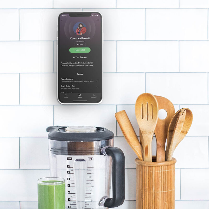 A smartphone mounted on a kitchen backsplash using a black PopSockets PopMount G4 SF. The phone is displaying a music app, and the mount securely holds the phone in an upright position on the tiled wall. Below the mount, there is a blender with a green smoothie and a wooden container holding kitchen utensils, indicating the practical use of the mount for hands-free access to recipes or entertainment while cooking.