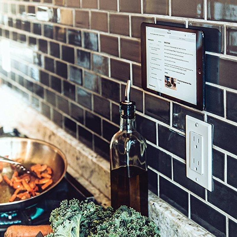 A tablet mounted on a kitchen backsplash using a PopSockets PopMount. The tablet is displaying a recipe, and the mount securely holds it in an upright position on the tiled wall. Below the mount, there is a bottle of cooking oil, vegetables, and a pan with food cooking on the stove, indicating the practical use of the mount for hands-free access to recipes while cooking. The setup showcases the convenience and functionality of the PopSockets PopMount in a kitchen setting.