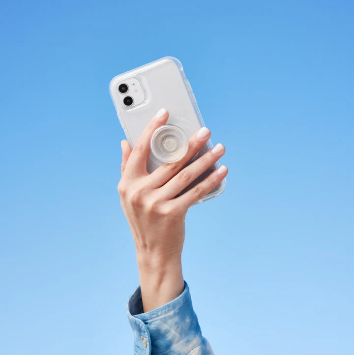 A hand holding a white smartphone with a clear case against a bright blue sky. The phone has a clear PopSockets PopGrip attached to the back. The grip is in its extended state, providing a secure hold. The clear design of both the case and the PopGrip allows the phone's original design to remain visible while offering functionality and style. The person is wearing a denim jacket, adding a casual touch to the scene.