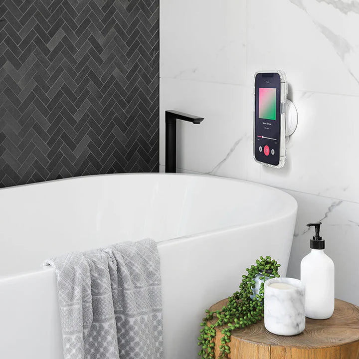 A smartphone is attached to a white tiled bathroom wall above a bathtub using a black PopSockets Suction Mount for MagSafe. The phone displays a music player app. The bathtub is modern and sleek, with a towel draped over the side and a black faucet. Nearby, there are bathroom essentials, including a soap dispenser and a small potted plant, creating a relaxing and functional bathroom setting