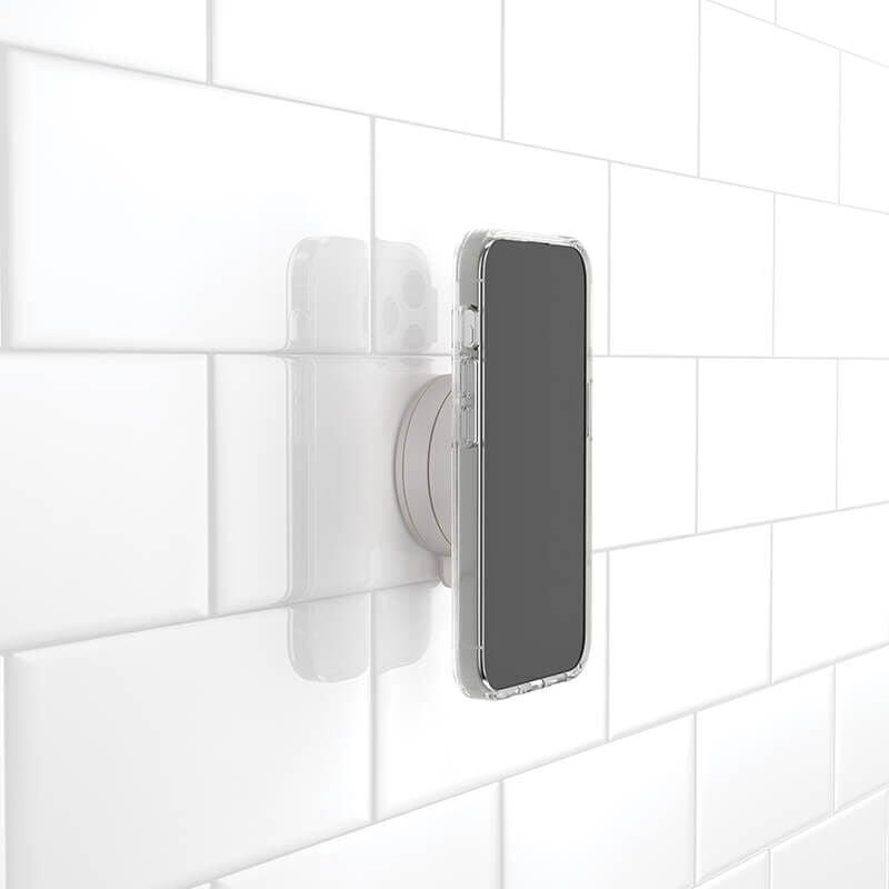 A smartphone is attached to a white tiled wall using a PopSockets Suction Mount for MagSafe in the color horchata. The mount securely holds the phone in place, demonstrating its ability to adhere to smooth, tiled surfaces. The phone is in a clear case, showcasing the practical application of the mount for hands-free use in various environments.