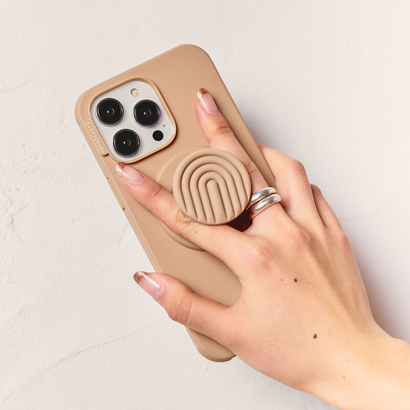 A hand with a manicure is holding a smartphone with a beige case and a PopOut Latte Curves PopGrip attached. The PopGrip features a latte-colored design with curved lines, providing a comfortable and stylish grip for the device