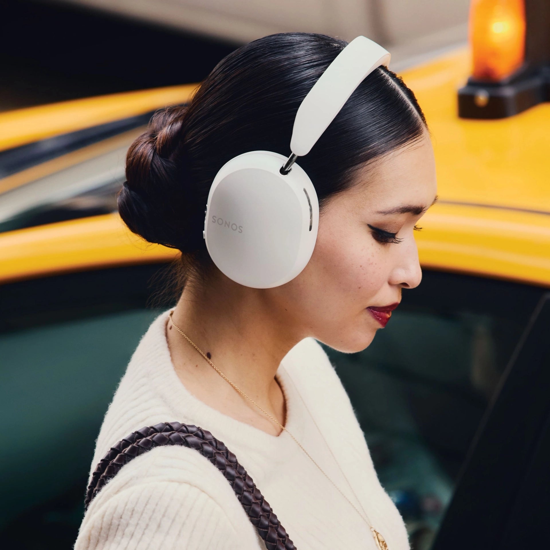 Woman wearing white Sonos wireless over-ear headphones, highlighting sleek design and comfort while standing near a yellow taxi.