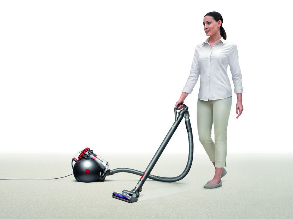 A modern canister vacuum cleaner being used on a soft carpeted surface, operated by a person dressed in a crisp, white button-up shirt and light-colored pants, showcasing efficient suction and maneuverability.