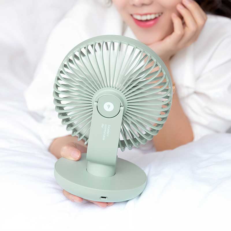 A pastel green portable desk fan held by a person lying on a bed with white bedding. The fan features a sleek circular blade design, a tilting mechanism, and a sturdy round base with a charging port. The person, smiling in the background, adds a cozy and relaxed vibe to the scene.