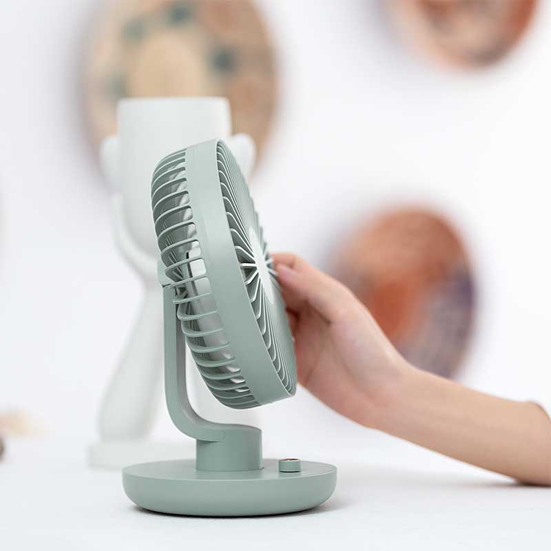 A small desk fan in a pastel green color with a minimalist design, featuring a circular blade enclosure and a sturdy round base with a single button. A hand is adjusting the fan's tilt angle. The background is softly blurred with abstract decor and muted tones, creating a clean and modern aesthetic.