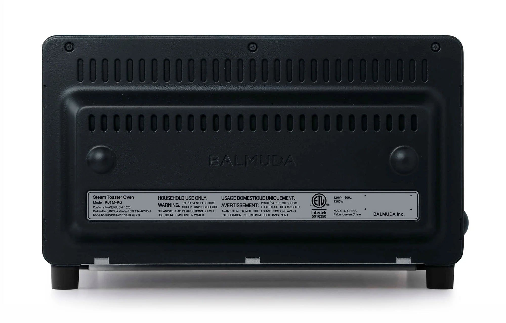  The image shows the back view of a Balmuda toaster in a matte black finish. The back panel features a series of horizontal ventilation slots near the top for airflow and cooling. The center of the panel has the brand name "BALMUDA" embossed into the surface. Below the brand name, there is a label displaying important safety and usage information, including warnings and certification details. 