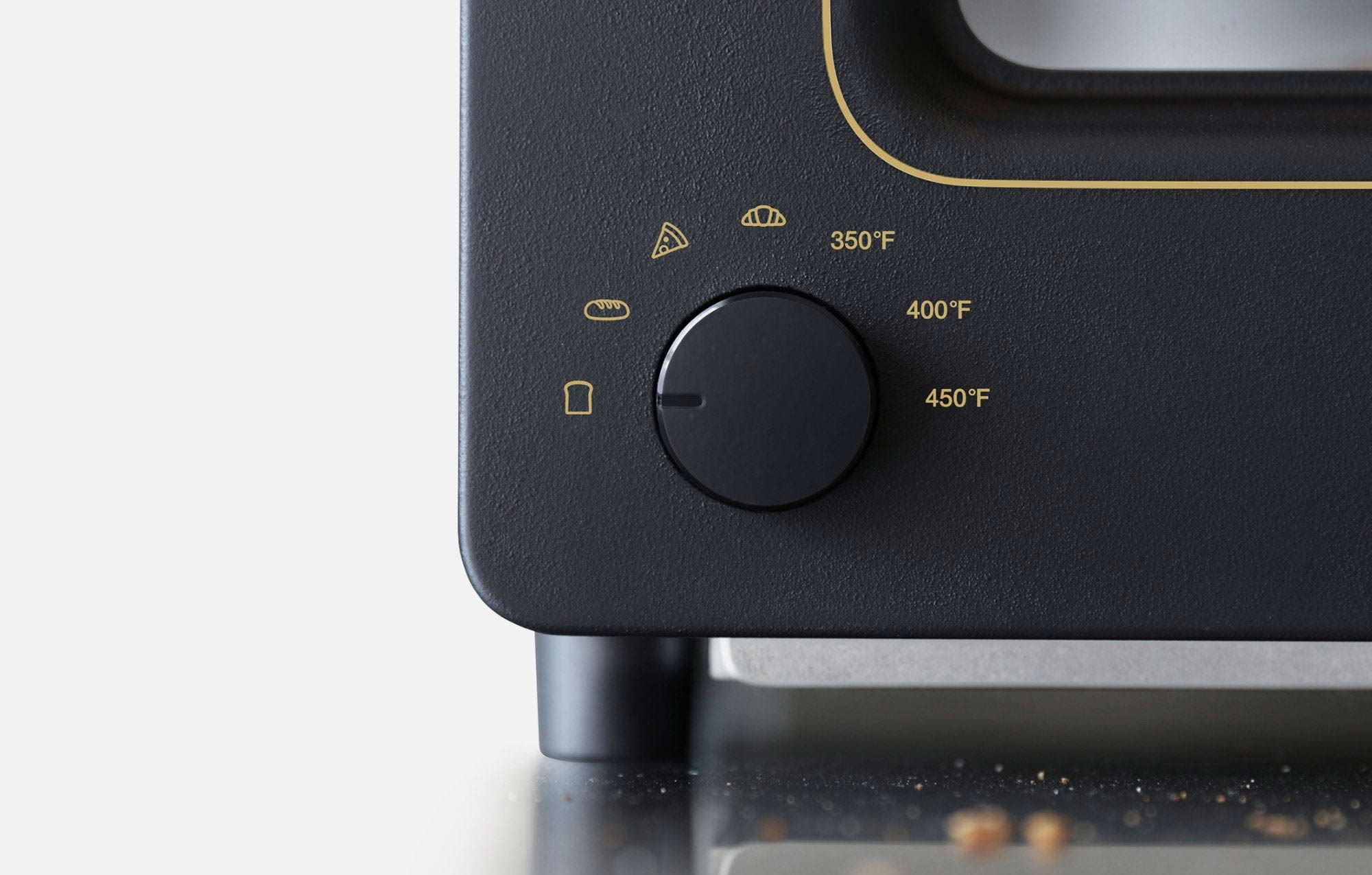  The image shows a close-up of the temperature control dial on the left side of a Balmuda toaster in a matte black finish. The dial is surrounded by icons representing different toasting modes: a slice of bread, a croissant, and a baguette, along with temperature settings of 350°F, 400°F, and 450°F. The dial is positioned on a sleek black surface, with subtle gold text and icons adding a touch of elegance. The surrounding area of the countertop shows a few breadcrumbs, indicating the toaster's use. The desi