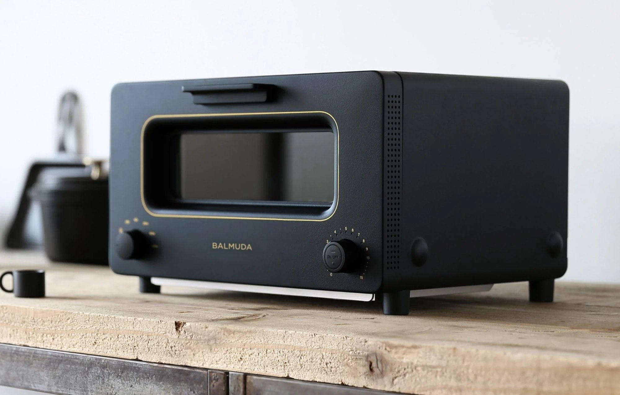  The image shows a side view of the Balmuda toaster in a matte black finish with gold accents, placed on a rustic wooden countertop. The toaster's design features a large rectangular viewing window, centered with the brand name "BALMUDA" in gold lettering beneath it. Two control dials are visible: one on the left for adjusting the temperature and another on the right for setting the timer. The setting emphasizes a minimalist, modern aesthetic, with the toaster's sleek design contrasting with the natural tex