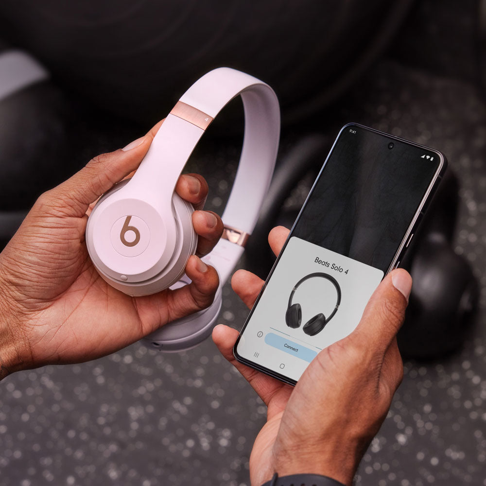 The image showcases a person holding a pair of Beats Solo 4 headphones in one hand and a smartphone in the other. The smartphone screen displays the Beats Solo 4 connected interface, indicating that the headphones are being paired with the device. The setting appears to be a fitness or gym environment, suggested by the background and the person’s active posture. The image highlights the ease of connectivity and the stylish design of the Beats Solo 4 headphones.