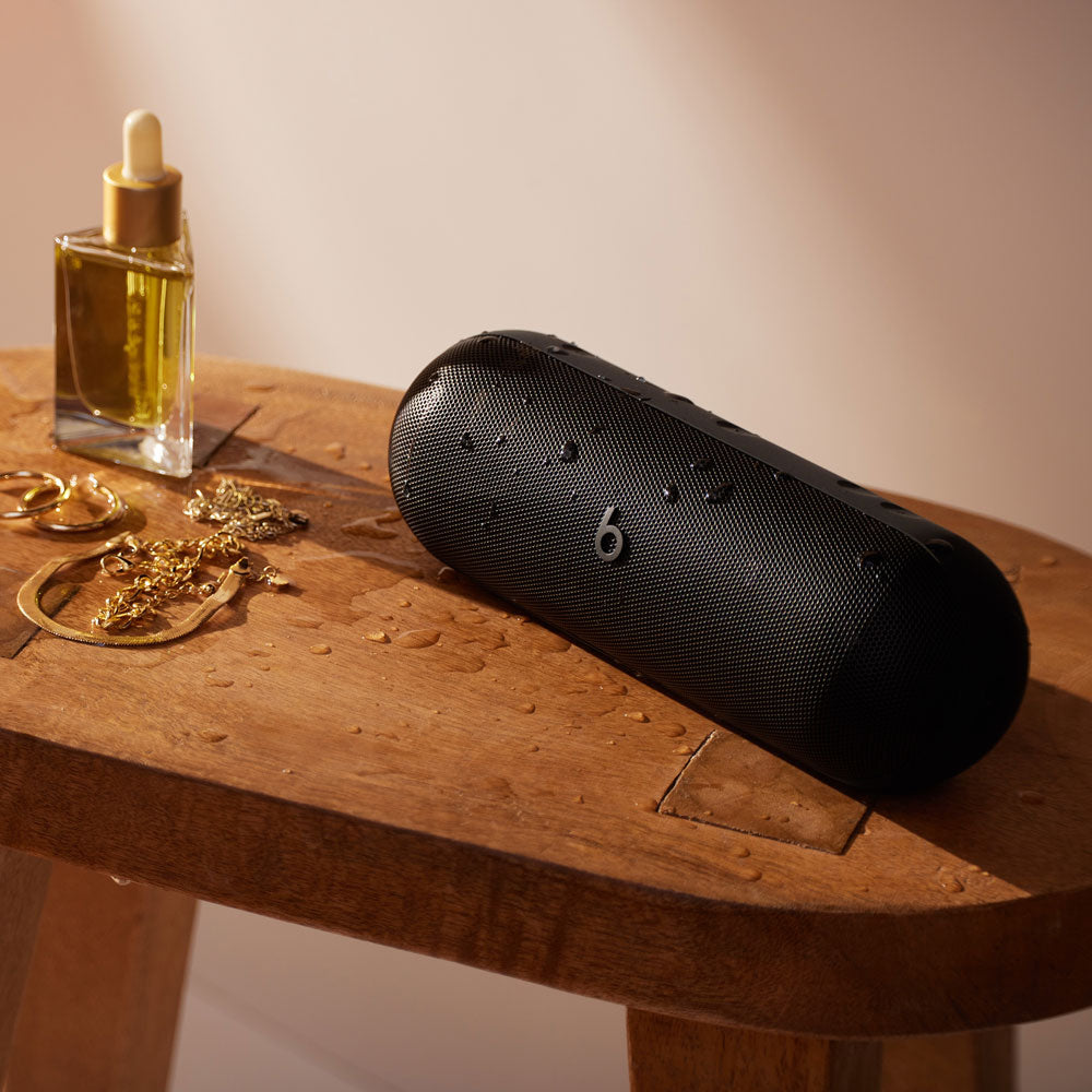 Alt text for this image:

A black Beats portable speaker with water droplets on its surface, sitting on a wooden table next to jewelry and a bottle of oil, highlighting its water-resistant feature and modern design in an elegant setting.