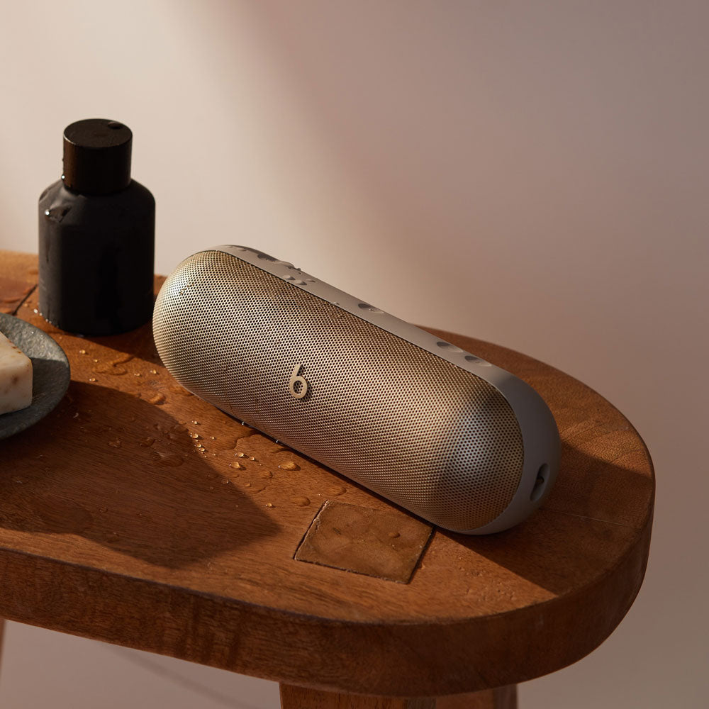 Alt text for this image:

A beige Beats portable speaker sitting on a wooden table, with a few water droplets nearby and a bottle of toiletries, highlighting its water-resistant design and suitability for bathroom or spa settings.