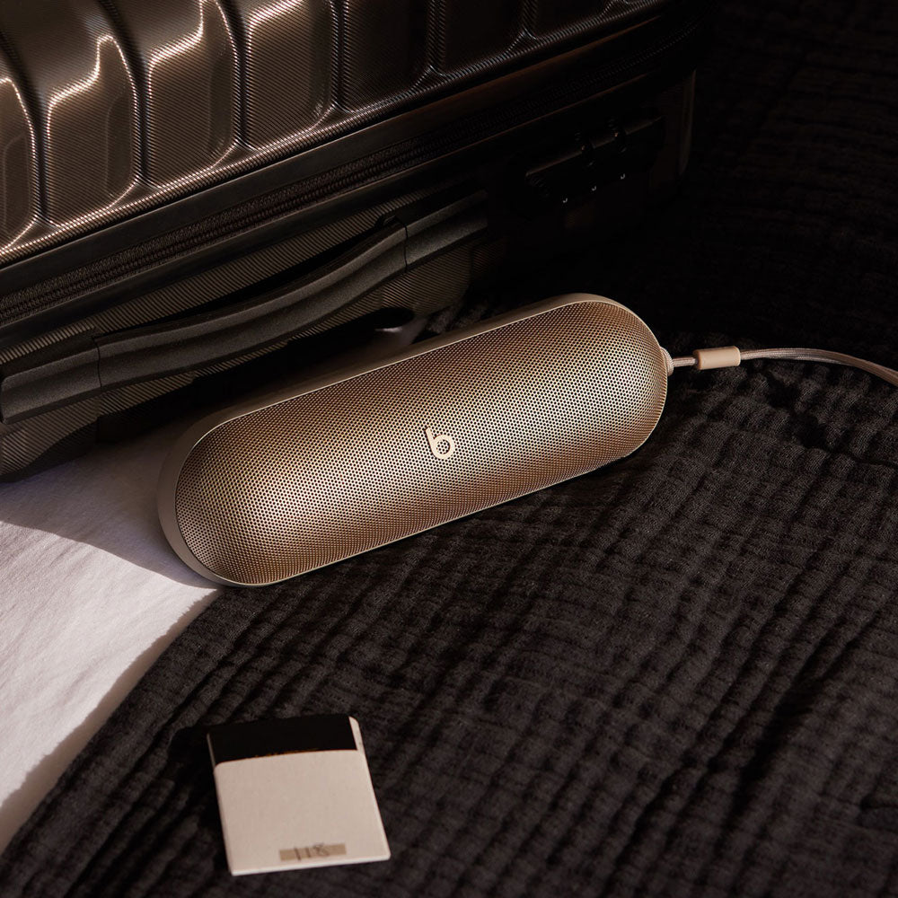 Alt text for this image:

A beige Beats portable speaker placed next to a travel suitcase on a soft black fabric, illustrating its portability and travel-friendly design, with a charging cable connected.