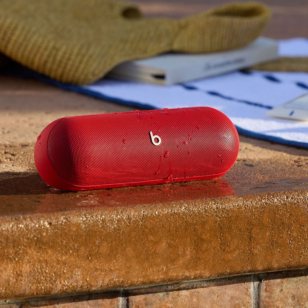 For the image you just uploaded, here is an alt text suggestion:

A red Beats portable speaker with water droplets on its surface, placed by a poolside, highlighting its water-resistant feature. A towel and a book in the background emphasize its suitability for outdoor use.