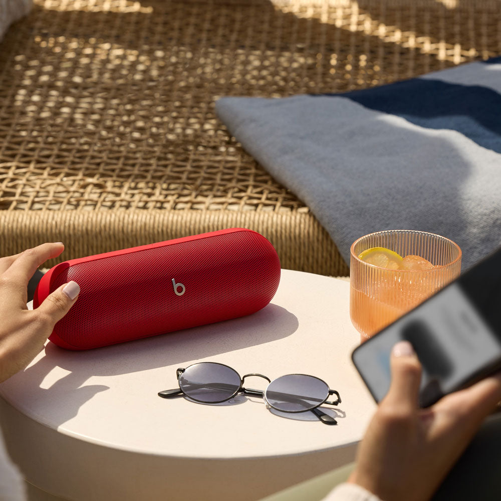 A red Beats portable speaker placed on a table outdoors, alongside sunglasses and a drink, creating a relaxed summer vibe.