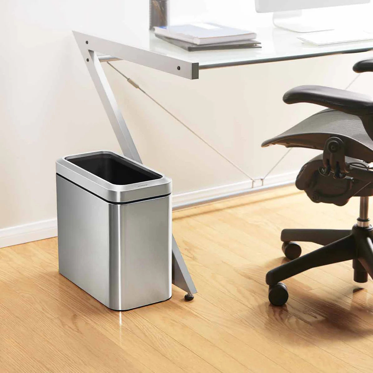 The image shows a modern office setting with a sleek, rectangular stainless steel trash can placed beside a glass-top desk. The trash can has a minimalist design with a polished finish and black accents around the top edge, which complements the contemporary workspace. The compact size fits neatly next to the desk, making it convenient for disposing of waste without taking up much space. The office chair, light wood flooring, and the overall clean and organized environment highlight the trash can's stylish 