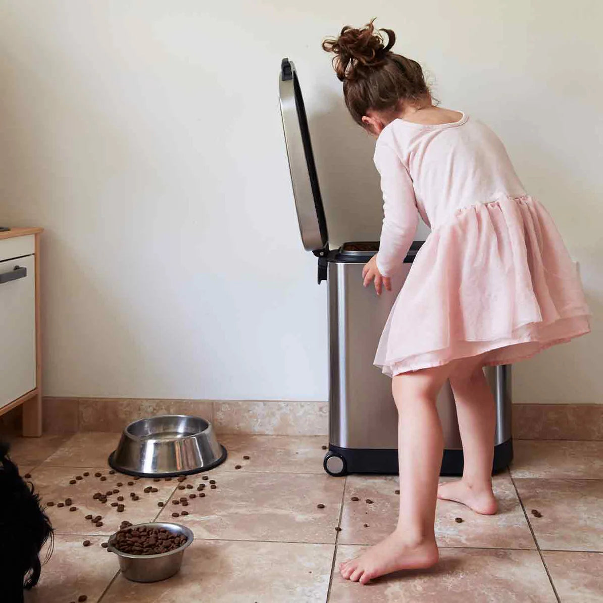 The image shows a young girl in a light pink dress reaching into a stainless steel trash can with a lifted lid in a kitchen or dining area. The trash can is sleek and modern, with a polished finish and a rectangular design. On the tiled floor beside the girl, dog food has spilled from a bowl, creating a slightly messy scene. A larger metal dog bowl is also visible nearby. The setting conveys a moment of interaction between the child and the trash can, possibly cleaning up after a pet. The image highlights t