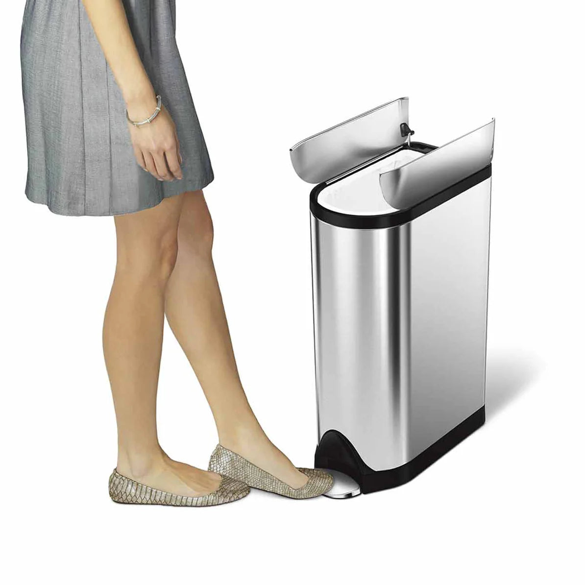 The image shows a person using a Simplehuman trash can. The can is sleek and modern, with a stainless steel exterior and a black trim. The lid is open, revealing the top of the trash can, which has a butterfly-style lid that splits in the middle and opens from the center. The person in the image is wearing a light gray dress and flat shoes, and is pressing a foot pedal at the base of the trash can, which is used to open the lid hands-free. This design is both stylish and functional, ideal for modern kitchen