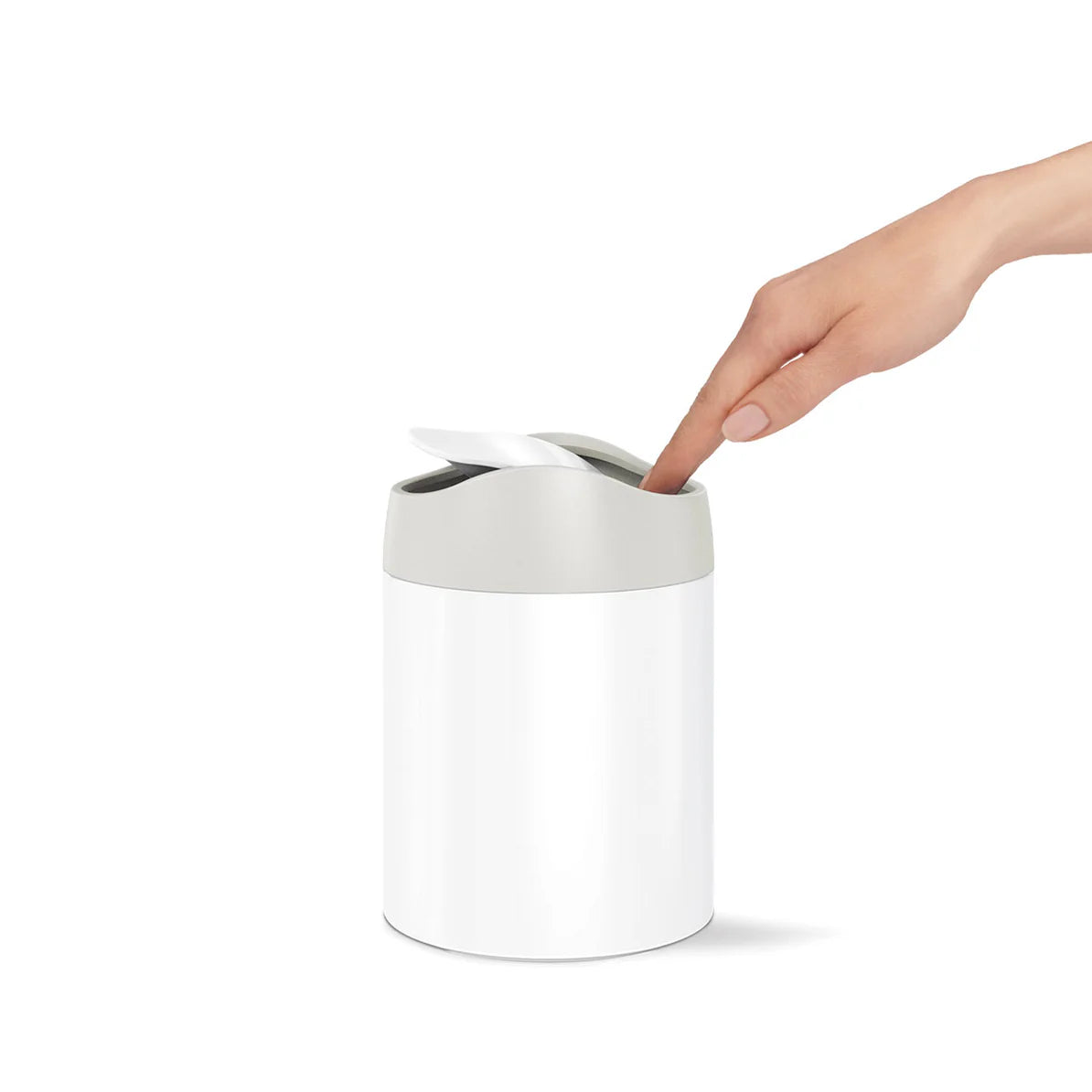 The image shows a Simplehuman mini round trash can with a white body and a matte gray swing lid. A person's hand is seen pressing down on the swing lid, demonstrating the ease of use. The trash can is compact and cylindrical, designed for small spaces like bathroom countertops, vanities, or desks. The white and gray color combination gives it a clean, modern appearance, making it a stylish and functional accessory for any contemporary setting. The swing lid is designed to open with a light touch and close a