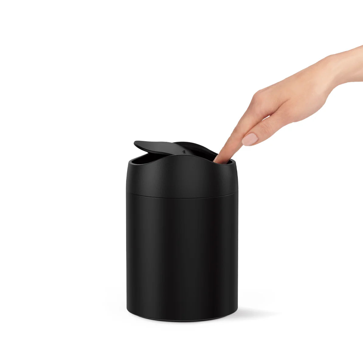 The image shows a Simplehuman mini round trash can with a swing lid, finished in matte black. A person's hand is shown pressing down on the swing lid, demonstrating the ease of use. The trash can is compact, making it ideal for small spaces such as bathroom countertops, vanities, or desks. The all-black finish gives the trash can a sleek, modern appearance, while the swing lid provides convenient and hygienic disposal of small items. This design combines practicality with a minimalist aesthetic, making it a