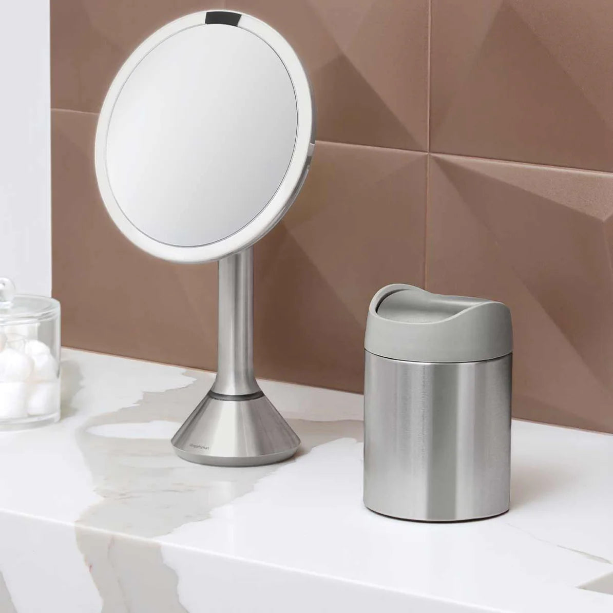 The image shows a modern bathroom vanity setup featuring two Simplehuman products. On the left is a Simplehuman sensor mirror with a brushed stainless steel finish and a sleek, circular design. The mirror is mounted on a tall, conical stand and is known for its LED lighting and sensor-activated features that provide optimal lighting for grooming tasks.