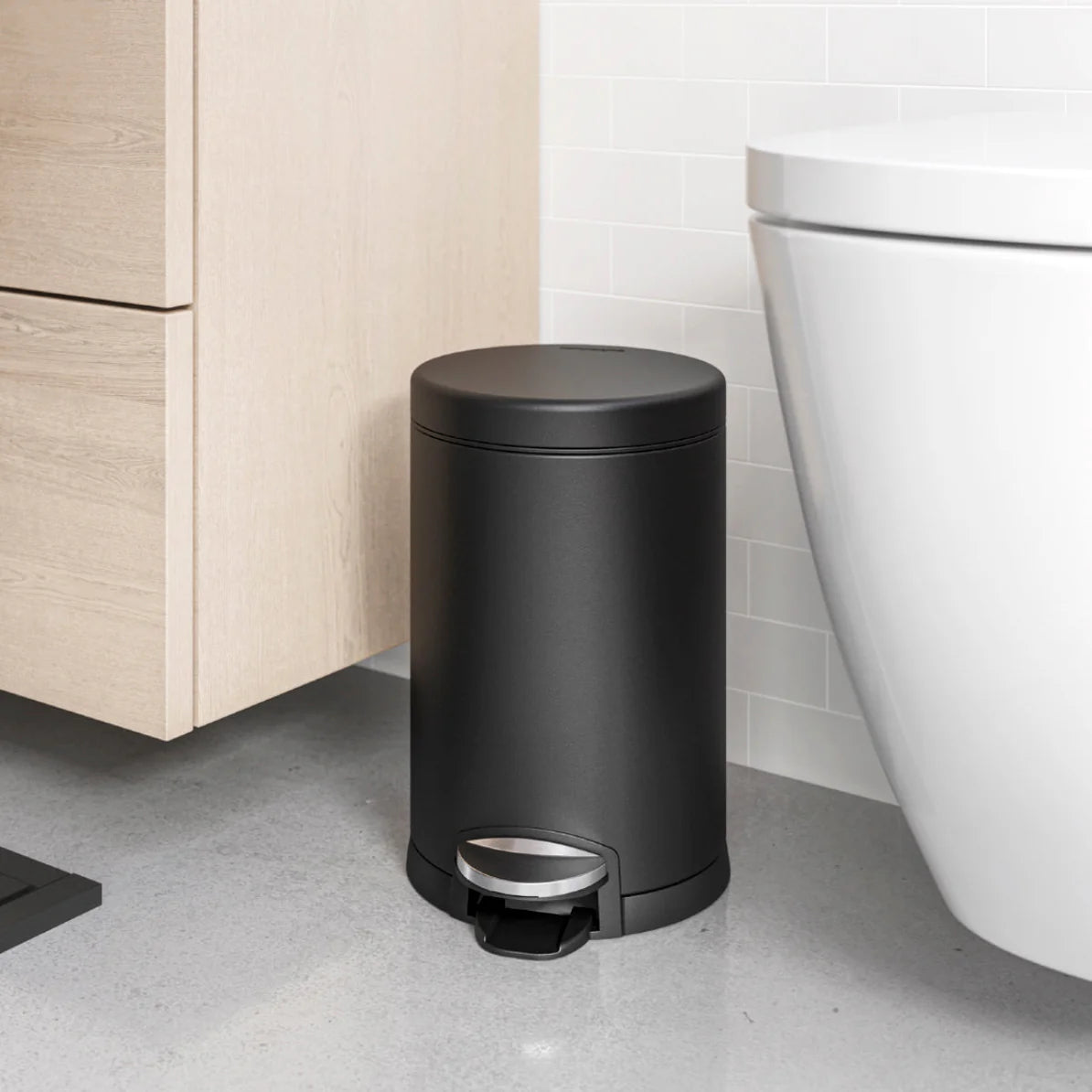 The image shows a sleek, black cylindrical trash can positioned in a modern bathroom setting. The trash can features a foot pedal for hands-free operation and has a minimalist design that complements the contemporary decor. It is placed near a wooden cabinet and next to a white toilet, blending seamlessly into the clean, uncluttered space. The matte black finish adds a touch of elegance and sophistication, making it a stylish yet functional addition to the bathroom.