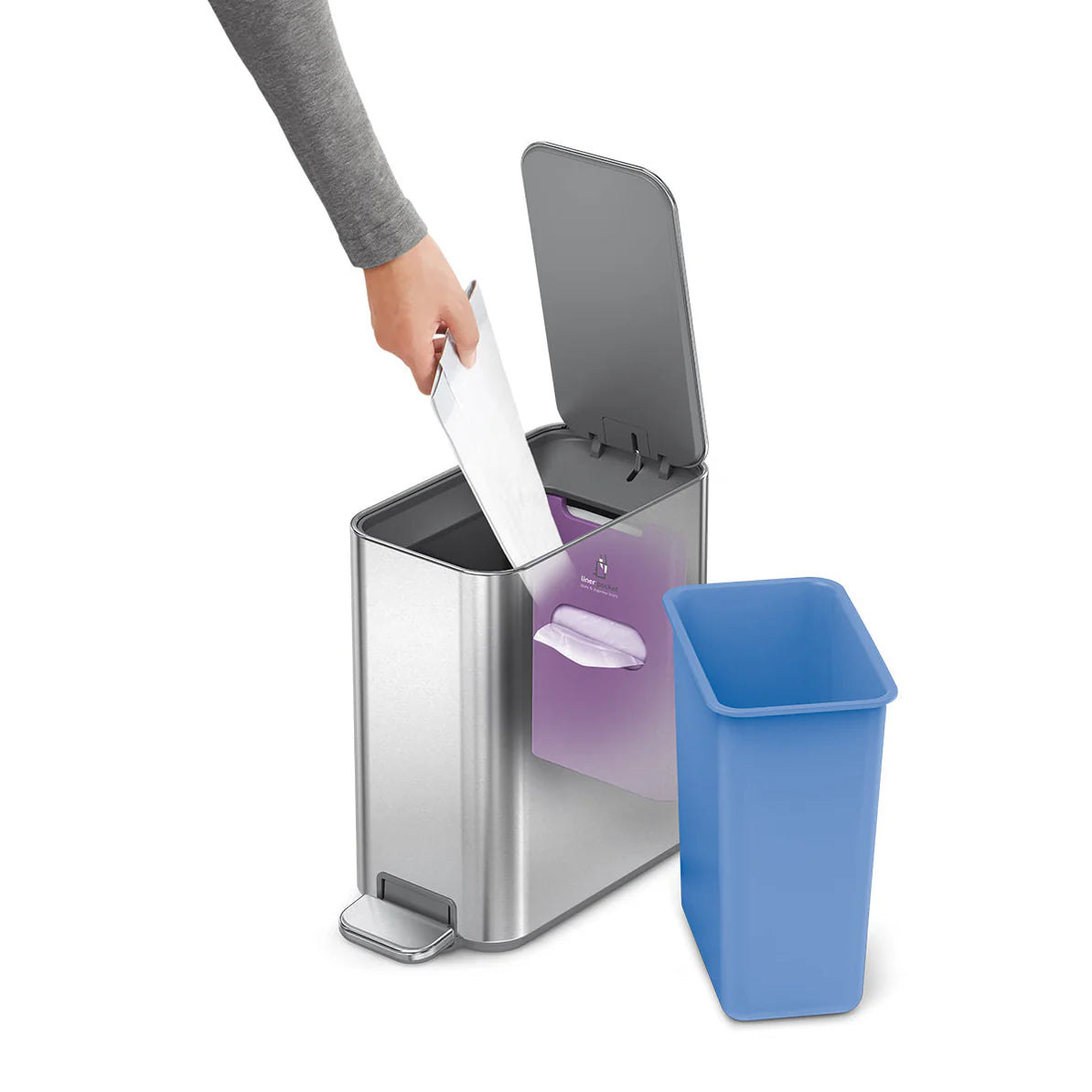 The image shows a stainless steel trash can with a rectangular design. The lid is open, and a person is seen placing a piece of trash into the can. The trash can features a blue inner bin that has been removed and placed next to the can, illustrating how it can be easily taken out for cleaning or replacing the trash bag. The can also has a foot pedal at the bottom front, which allows for hands-free operation. The overall design is sleek and functional, with the stainless steel exterior providing a modern an