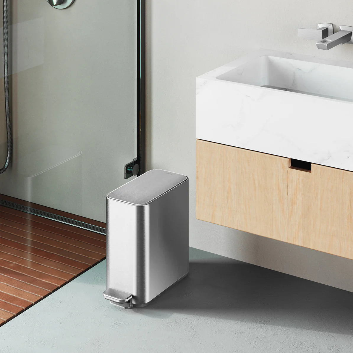 The image shows a modern bathroom setting with a sleek, stainless steel trash can placed next to a floating wooden vanity with a white countertop and sink. The trash can has a rectangular shape with rounded edges and is equipped with a foot pedal at the front for hands-free operation. The bathroom features a glass shower door with a wooden floor inside the shower area, adding a warm contrast to the minimalist design. The stainless steel trash can complements the contemporary aesthetic of the bathroom, provi